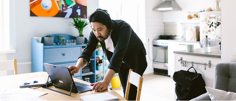A man wearing headphones leans over a table in a home kitchen to work on a tablet.
